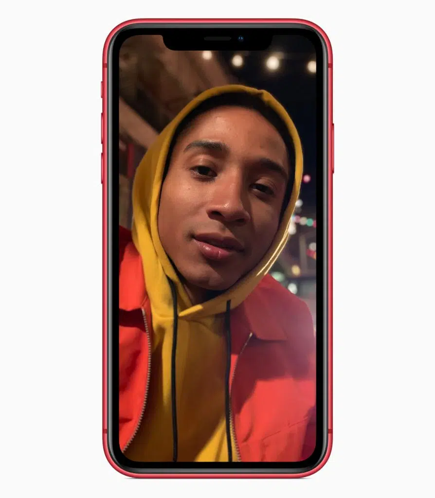 Imagem mostra um iphone xr vermelho exibindo a foto de um jovem negro com a cabeça coberta pelo capuz de um moletom amarelo mostarda e jaqueta vermelha sobreposta. A foto foi tirada usando modo retrato, por isso o fundo está desfocado.