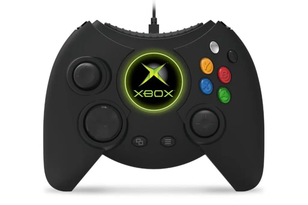 O controle duke está em pé com um fundo branco. O controle é revestido de preto e os botões ao lado direito contém algumas cores, como azul e vermelho. No centro, há o logo da xbox com destaque em verde.