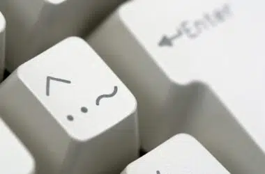 Detalhe de teclas de um teclado bege. Em destaque, tecla com acento circunflexo no canto superior esquerdo. Logo abaixo, à esquerda, dois pontos e à direita, um til.