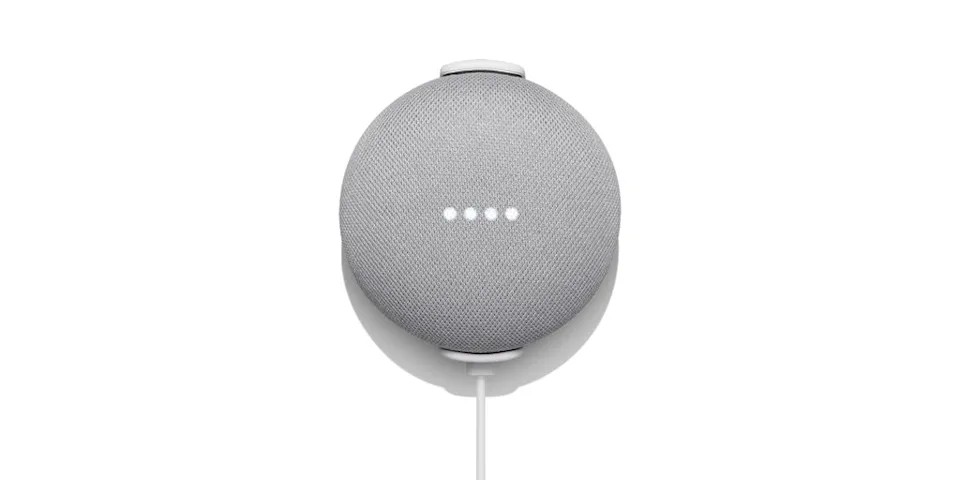 Parede branca e nest hub mini no meio na cor cinza, 4 pontos de luz no meio indicando que está ligado e um cabo de energia conectado no aparelho.