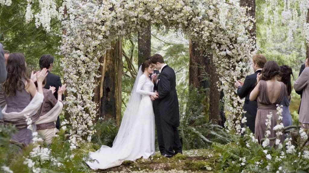 Na foto está o altar do casamento de bella e edward. Os protagonistas estão ao centro da foto, acima de suas cabeças há um arco de flores brancas. Convidados assistem a cerimônia e aplaudem.