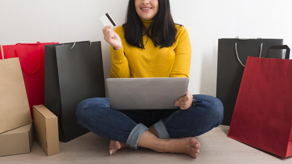 Mulher ao centro da imagem com um notebook e cartão de crédito em mãos se prepara para fazer compras internacionais online.