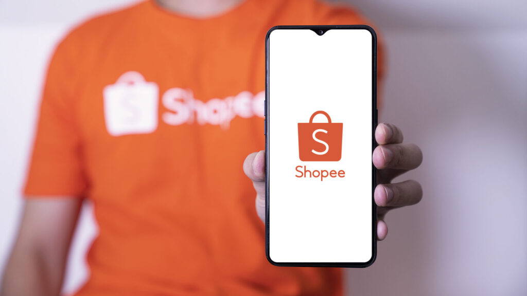 Na foto há um homem segurando um aparelho celular, nele é possível ver o símbolo da shopee.