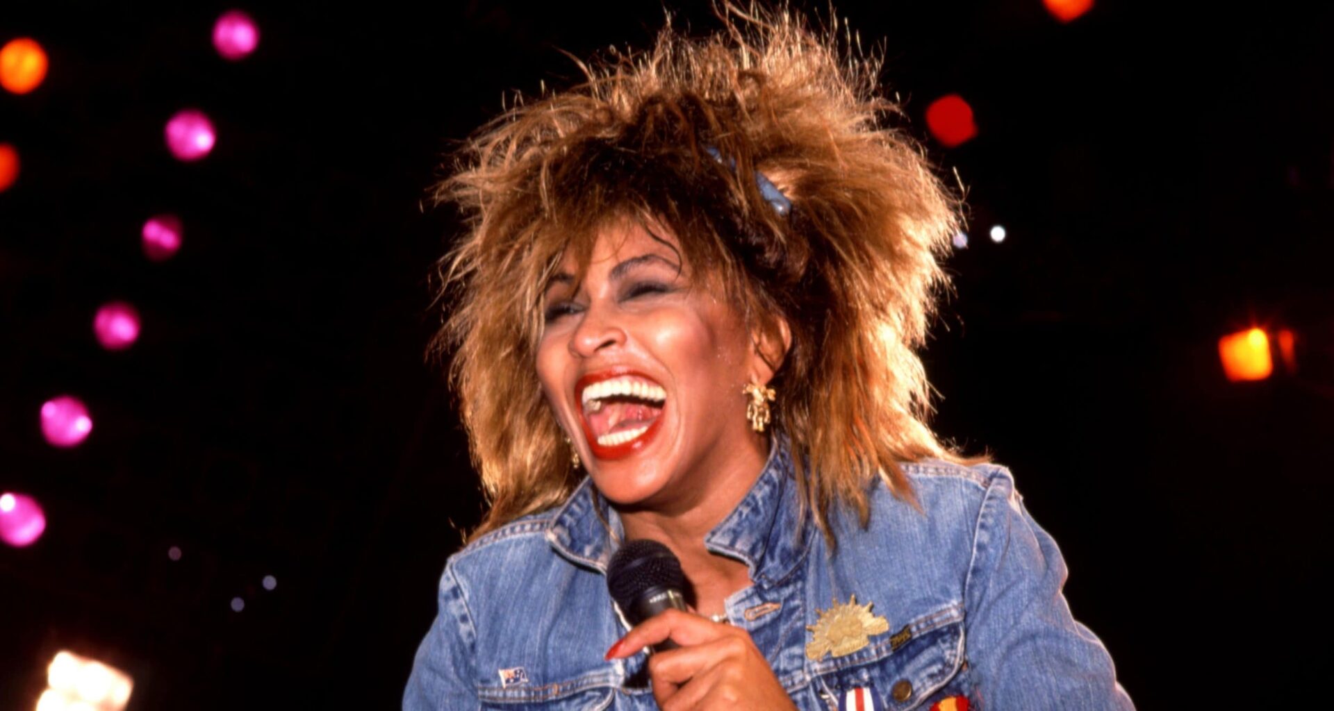 Tina turner t alt | destaq | cantora vista sob iluminação forte, vestindo jaqueta jeans, segurando microfone contra o peito e sorrindo de boca bem aberta