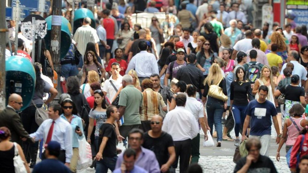 Censo 2022 t alt pessoas na rua, vestindo diferentes roupas cobrindo todo o corpo, com alguns orelhões em cor verde à dir.