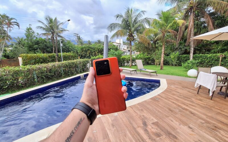 Homem segurando smartphone laranja em cenário com piscina, coqueiros e céu azul